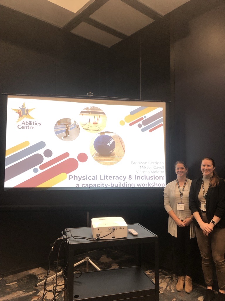 Two people stand next to a large presentation screen before presenting the Physical Literacy & Inclusion workshop.
