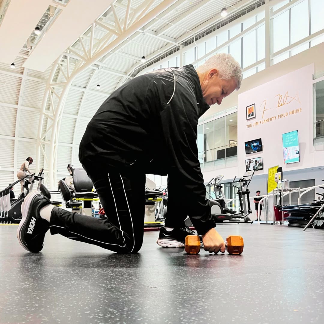 An older gentleman kneels as he prepares to lift a weight.
