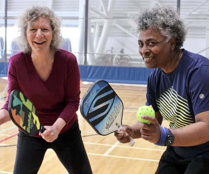 Two people smiling and ready to play pickleball as they hold up their paddles in the ready position.